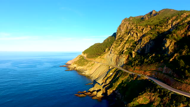 aerial drone shot of a Road on the cliff side between the mountain and the sea at sunset