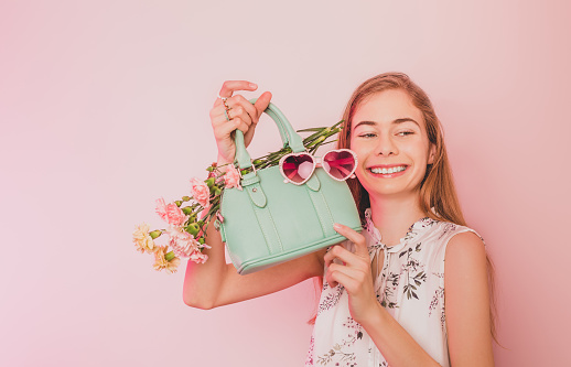 Fashion spring accessories. Happy smiling teenager girl holding mint green handbag (purse), heart shaped sunglasses and flowers on pastel pink background. Free copy (text) space.