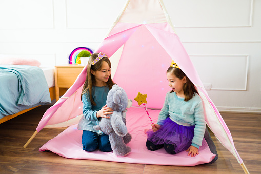 Beautiful little girls using toys teddy bears while playing dressed as princesses inside a cute pink teepee