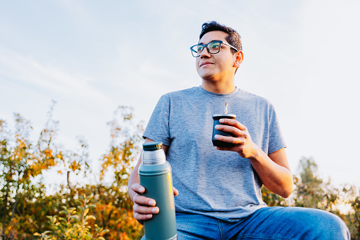 Hispanic young man drinking mate and relaxing, in a rural space, on top of an old tractor.