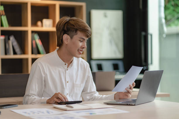 istock Serious young asian man working in office at desk. Holding and looking at document. Graph. Planning in office. calculation report Using laptop and calculator, online meeting concept 1460943833