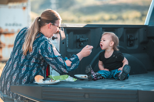 Adorable small child being fed by mother, The kid is seated in the back of a truck and is having his mouth wide open.