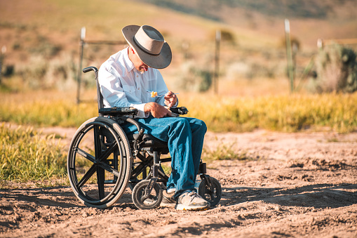 Full length shot of white adult man in a wheelchair. He is seated while eating, he wears jeans and a cowboy hat.