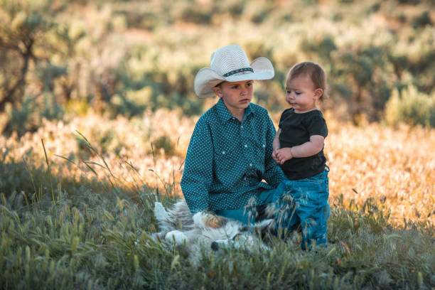 Full length shot of little male kid and bother playing and petting a white dog in the field. The oldest sibling is wearing a cowboy hat.