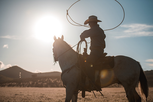 Candid shot of mid adult man with a roping lasso. Silhouette of a cowboy twirling a lasso around while the sun is up on the sky.