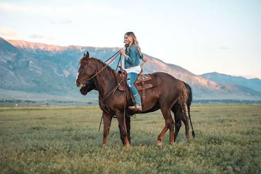 Full length shot of cowgirl horseback riding in the large field. She is having fun in the countryside while looking away.