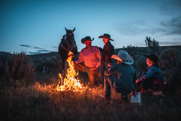 Loving father and sons smiling and taking a rest next to a bonfire. They are all wearing cowboy hats and there is a horse next to them.