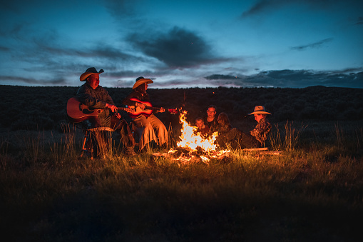 Outdoor shot during the late afternoon. Happy family of western americans gathering next to a campfire. Two are playing the guitar and the rest are looking at the fire.