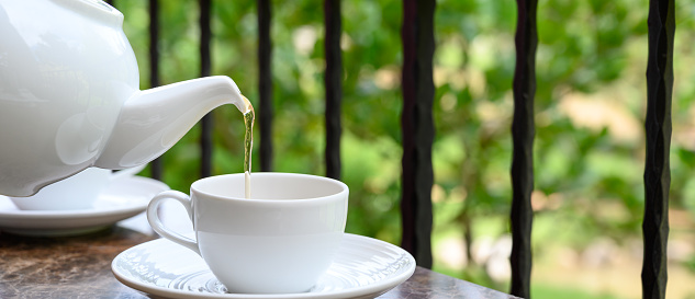 Pouring hot tea from white ceramic teapot into porcelain cup. Outdoor breakfast on balcony cafe