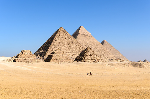 Pyramids of Giza, Giza Necropolis, Egypt - January 22, 2022: Man walking with camels at the Pyramids of Giza. \n\nThe Giza Necropolis is a large area near Giza, a suburb of Cairo that has many tombs and burial sites. It is also where you can find the famous Pyramids of Giza, the Great Sphinx, and more. The name comes from the ancient Greek word nekropolis, which literally means “city of the dead”.\n\nThe Giza Complex was once near the ancient Egyptian capital of Memphis, and the Giza plateau is where a number of pharaohs, queens, and nobles of the Fourth Dynasty of ancient Egypt were buried. \n\nAll three of Giza's pyramids and their elaborate burial complexes were built  from roughly 2550 to 2490 B.C. The pyramids were built by Pharaohs Khufu, Khafre, and Menkaure.