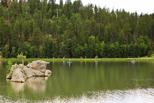 Hikers and paddle boarders on and along Sylvan Lake with a hill of green trees in the background in a summer South Dakota landscape
