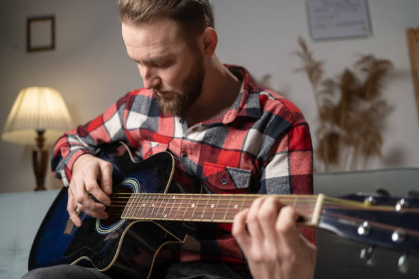 istock Focused serious man playing the guitar while sitting on sofa at home in the evening. Enjoying carefree time. 1460953180