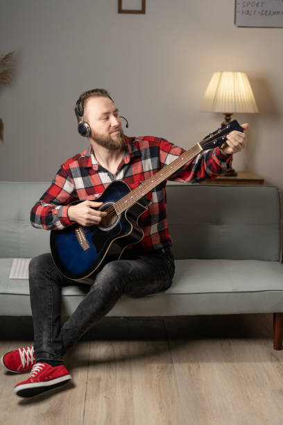 istock Young man playing guitar while sitting on sofa in living room in the evening. 1460953246
