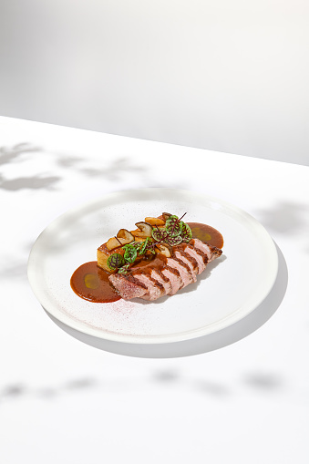 Roasted duck breast with potato gratin and sauce on light background. Fried duck fillet on white plate with hard shadows. Elegant summer menu. French cuisine - duck breast with gravy