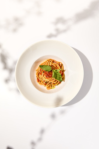 Vegetarian pasta with basil and tomatoes on white plate. Tomato pasta in summer menu with hard shadows. Spaghetti with tomatoes on light background. Vegan food - spaghetti marinara