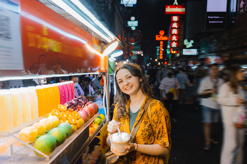 Young Caucasian woman eating coconut ice cream  and looking at camera on night market in Chinatown, Bangkok