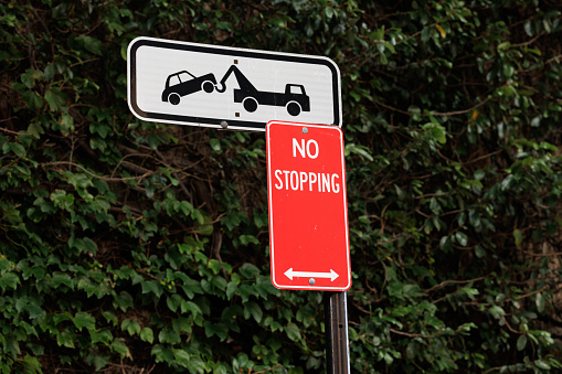 Red metal No stopping sign on a metal pole, with a planted wall behind. Above is a warning that cars will be towed away.