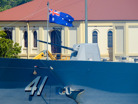 The Australian flag flies on the bow of HMAS Hobart, while the turret belongs to HMAS Brisbane, docked side-by-side.  HMAS Brisbane (left) and HMAS Hobart (right) are two of three Hobart Class guided missile destroyers of the Royal Australian Navy.  They are docked at Garden Island, Sydney Harbour.  The building in the background has stained-glass windows.  This image was taken from Mrs Macquarie's Chair on a sunny and windy afternoon on 28 January 2023.