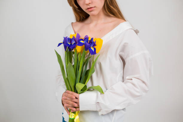ein blondes mädchen in weißem hemd und blauen jeans hält einen strauß tulpen und iris auf weißem hintergrund sitzend. 8. märz. blumenstrauß. junge frau mit blumen in nahaufnahme. strauß von tulpen und iris. frauenurlaub. frauentag. muttertag. - tulip blue close up cut flowers stock-fotos und bilder