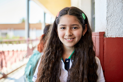 Head and shoulders view of Hispanic child with long wavy hair wearing private school uniform and standing outside classroom smiling at camera.