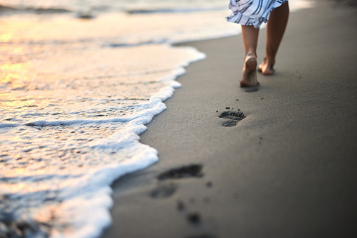 Back view of unrecognizable woman leaving footsteps in sand while walking on the beach at sunset. Copy space.