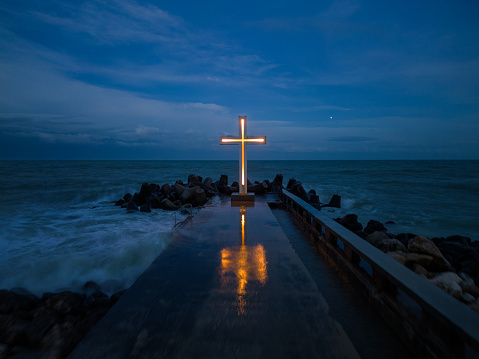 christian cross standing on pier in the stormy sea or ocean with dramatic sky at night