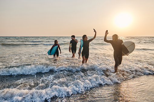 Cheerful couple of surfers giving each other high-five while leaving the sea after successful surfing at sunset. Their friends are in the background. Copy space.