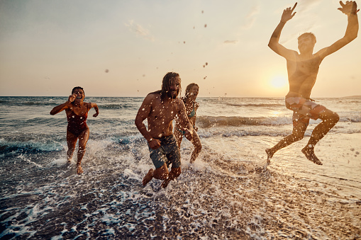 Group of cheerful friends having fun while running out of sea at sunset. Copy space.