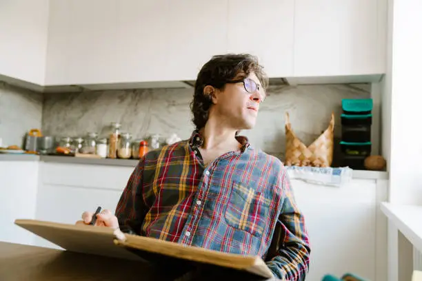 White man wearing glasses writing down notes while sitting in kitchen at home