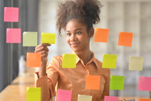 istock Smiling African American female manager putting sticker notes to share idea for startup project on glass board in office. 1461011471