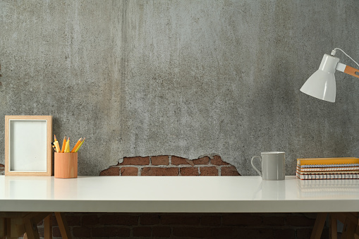 Stylish workplace with picture frame, books, pencil holder and lamp on wooden table against concrete wall. Copy space for your text.