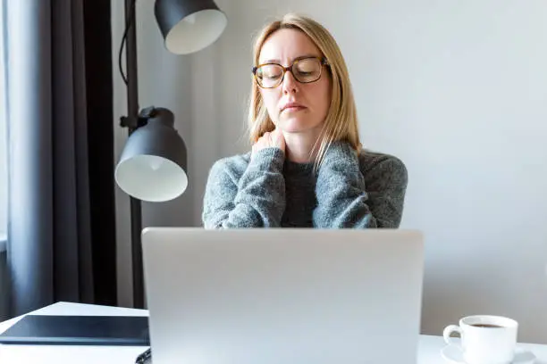 Tired female freelancer massaging neck in front of the laptop.