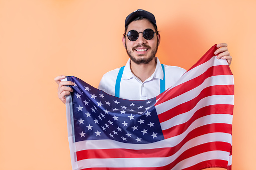 The happy young hipster holding USA flag, on light orange background.