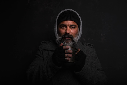 A homeless middle-aged man with a gray beard holding a cup of hot tea on a black background