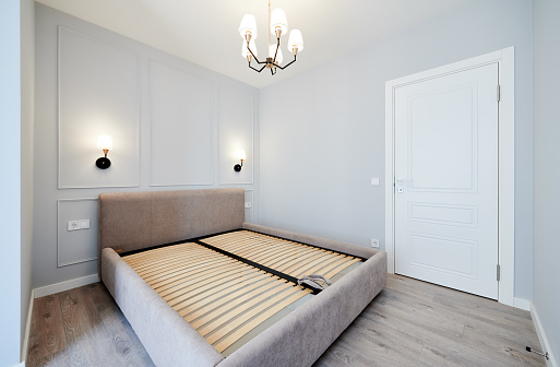 Bedroom with large bed, laminate floor, white walls, closed door and lamps in apartment after renovation. Interior of modern apartment with elegant minimalist design and comfortable sofa bed.