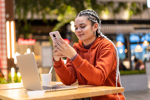 A cheerful pretty Caucasian lady with braids using her phone sitting in front of a laptop