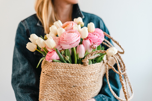 Woman holds a bouquet of white tulips and pastel pink and peach ranunculus in a jute bag.