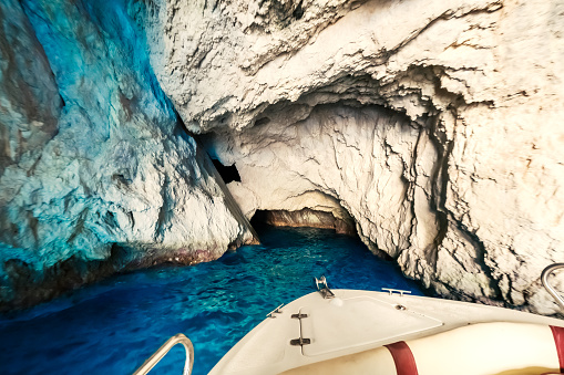 Zakynthos, Greece - July 16th 2022: View into a small sea cave, surrounded by beautiful blue water that seems to literally glow. This effect is achieved by the sunlight reflecting off the seabed. At the bottom of the picture you can see the bow of the small motorboat that took us into the grotto. No people visible.
