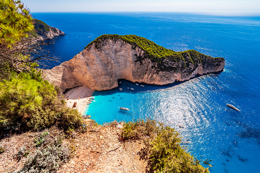 Zakynthos, Greece - July 16th 2022: View of the old, rusty shipwreck from the famous beach of Navagio, which has been named the world's best beach several times. The photo was taken from a vantage point on the edge of the cliffs, at peak times you sometimes have to queue for over an hour to catch a glimpse of the beach from this point. Some tourists cavort on the beach and marvel at the shipwreck, while the boats, which have unloaded these tourists are now waiting for a sign to pick them up again and anchor off the coast in the turquoise sea.