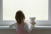 Little girl holding white teddy bear and standing alone at window and looking out from home. Back view. Waiting concept. Close up.