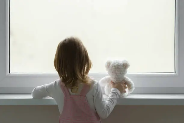 Photo of Little girl holding white teddy bear and standing alone at window and looking out from home. Back view. Waiting concept. Close up.
