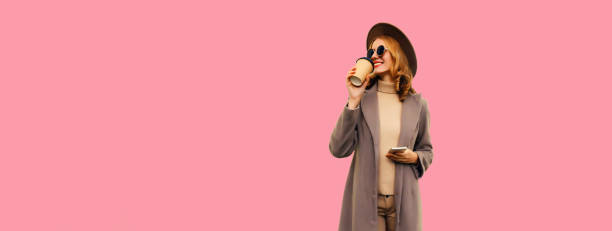 Portrait of stylish beautiful young woman model with cup of coffee and phone looking away wearing brown round hat and coat on pink background, blank copy space for advertising text stock photo