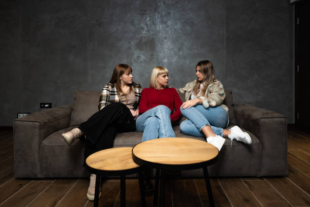 drei junge freundinnen sitzen zu hause auf dem sofa und tratschen über andere frauen und menschen, reden über ihr leben und machen witze übereinander. - rudeness whispering gossip humor stock-fotos und bilder