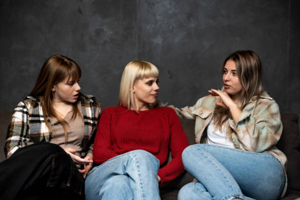 três jovens amigas estão sentadas no sofá de casa fofocando sobre outras mulheres e pessoas, falando sobre suas vidas e fazendo piadas umas sobre as outras. - rudeness whispering gossip humor - fotografias e filmes do acervo