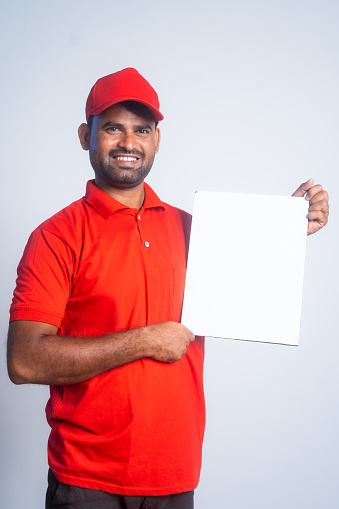 Vertical shot of happy painter standing with showing white empty board by pointing finger while looking at camera at house - concept of paint advertisement, employment and renovation