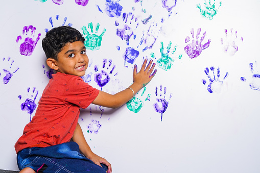 Kid creating mess by handprint paint on white wall by looking at camera at home - concept of mischief, playfulness and holidays