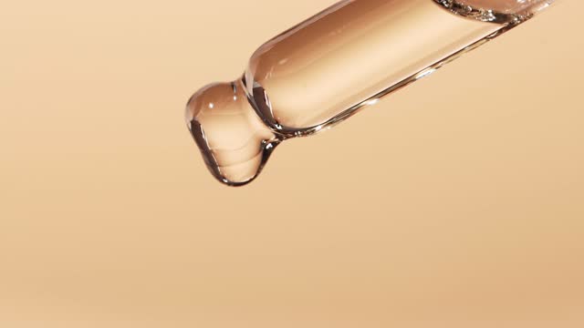 Essential oil dripping from a glass dropper, beige background, close-up. The concept of dermatology and cosmetology.