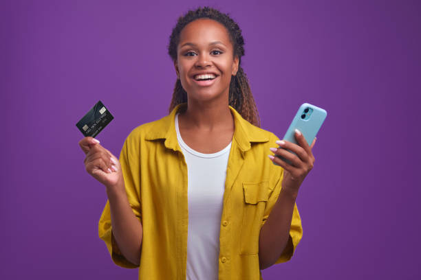 jeune femme afro-américaine souriante avec carte de crédit et supports téléphoniques dans le studio - internet e commerce credit card retail photos et images de collection