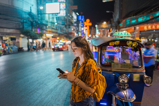 Young Caucasian woman with smartphone standing near tuk-tuk  in Chinatown in Bangkok