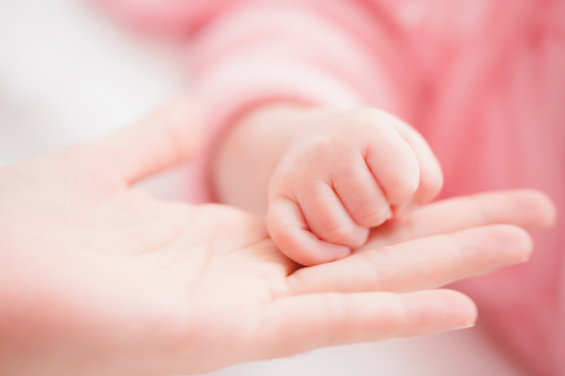 Close up mother holding hands newborn girl in a room. Adorable infant rests on white bedsheets, staring at camera looking peaceful. Infancy, healthcare and paediatrics, babyhood concept.